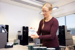 Etualalla on CD-levyjä neljässä eri pinossa. Henkilö, jolla on viininpunainen paita, pakkaa pöydän ääressä CD-äänikirjaa koteloon. Taustalla CD-äänikirjojen tekemiseen käytettäviä laitteita.