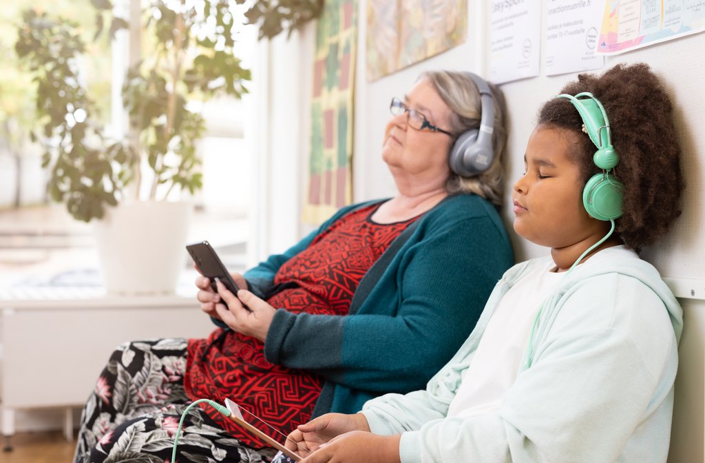 A child and an elderly person sitting next to each other on the floor leaning against the wall. Both are listening to an audiobook from Accessibility Library Celia with headphones. 