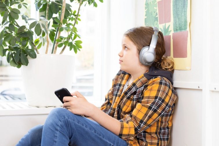 A child is sitting leaning against a wall and listening to an audiobook from Accessibility Library Celia on a mobile phone with headphones on. There is a house plant in the background.