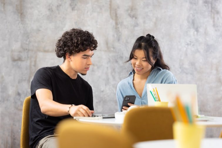 Two people are sitting at a round table with computers in front of them. They are looking at a mobile phone together, the other one has earbuds in their ears. The person holding the mobile phone is smiling. There are pens on the table.