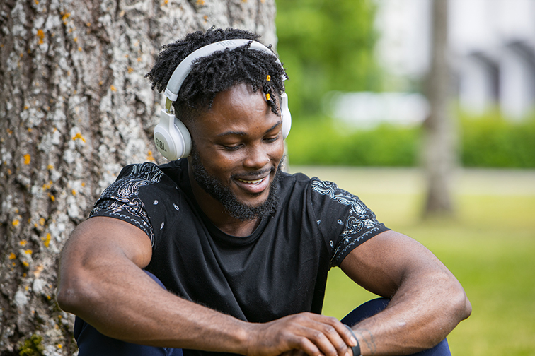 A person is sitting outside in front of a tree with his eyes directed downwards and his forearms resting on his knees. He is wearing headphones.