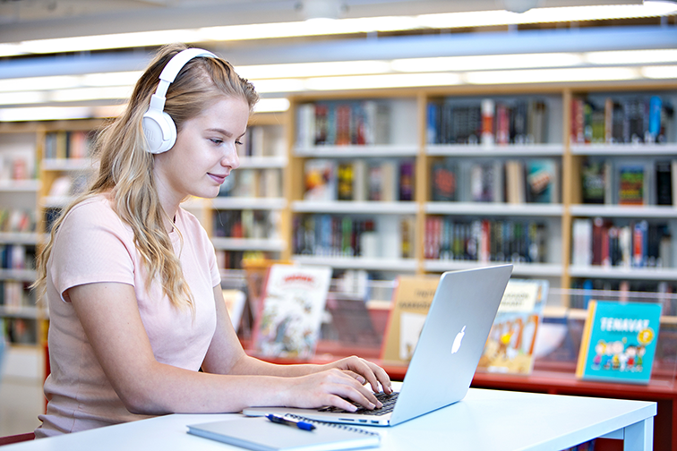 A young person is sitting in front of a computer in the library with headphones on. Bookshelves in the background. On the table a pen and a notebook. Picture of Accessibility Library Celia.