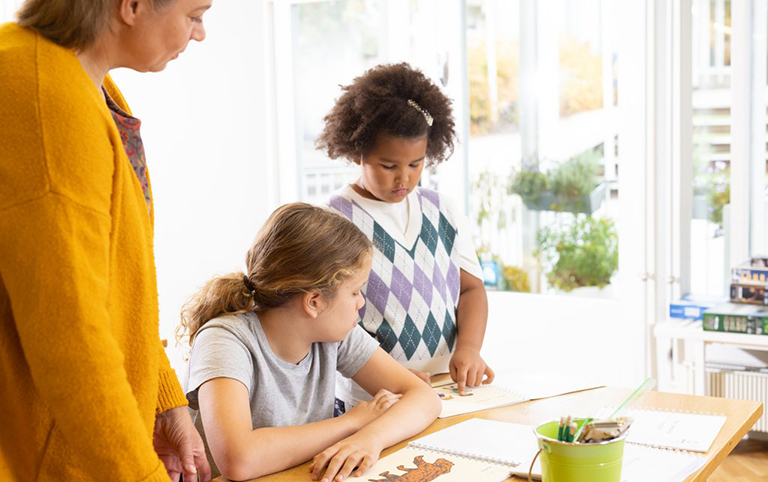 An adult and two children at a table. One child is reading a Braille book, the others are watching. On the table are tactile books and school supplies. Picture of Accessibility Library Celia.