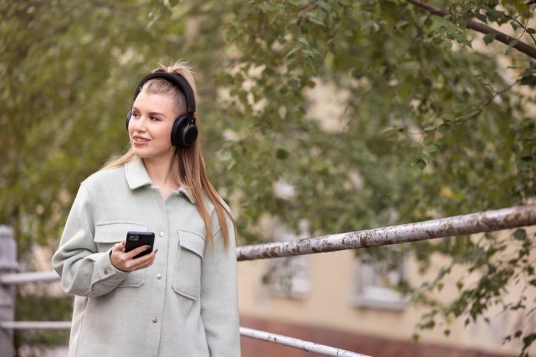 En person med hörlurar på promenerar utomhus och håller en telefon i handen. Bredvid personen finns ett räcke och i bakgrunden syns ett hus och ett träd med gröna löv.