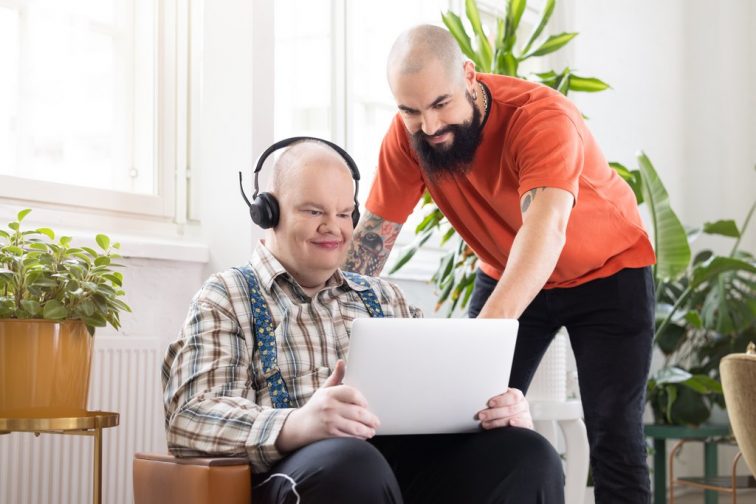 En person sitter i en stol framför fönstret med en bärbar dator öppen i knät och hörlurar i öronen. En annan person står bredvid hen, böjd framat för att titta på datorskärmen. I bakgrunden finns gröna växter.