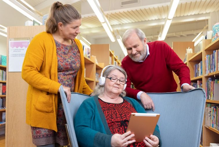 An elderly person sitting in an armchair in a library and listening to a talking book with a tablet in their hand and headphones on their head. Two people stand behind the chair. One is watching the other, another elderly person, who in turn is looking at the device. There are several bookshelves in the background.