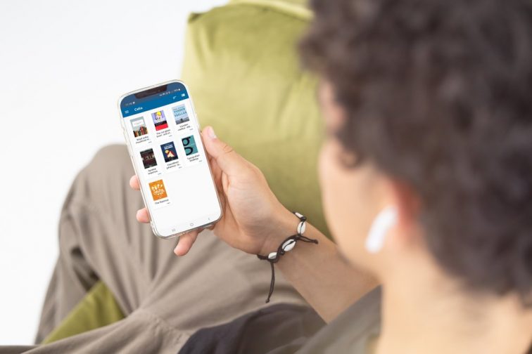 Over-the-shoulder shot of a person sitting and wearing white in-ear headphones. They are holding a phone with Celia’s Pratsam Reader talking book listening app open on the screen.