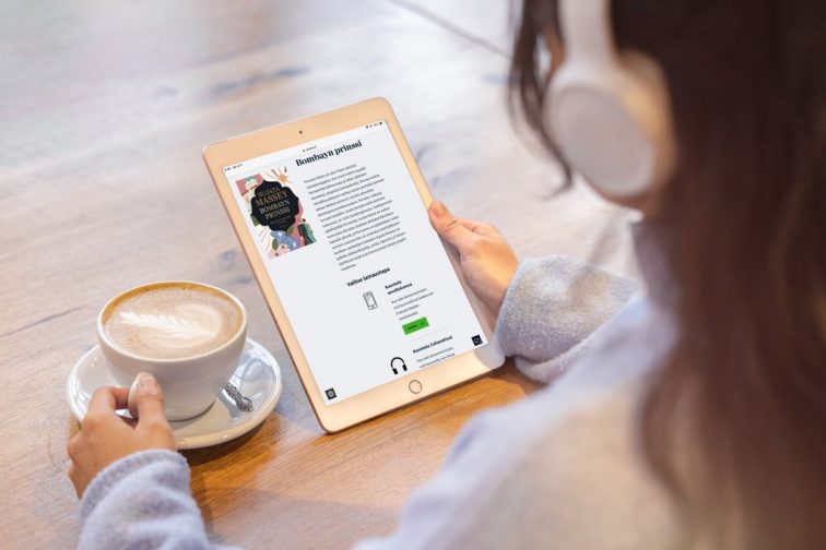 Over-the-shoulder view of a person wearing headphones. They are sitting at a table and holding a tablet with Accessibility Library Celia’s Celianet talking book service website open. On the table is a cup of coffee.